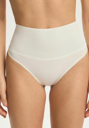 Kobieta nosząca białą bezszwową bieliznę z wysokim stanem, stojąca z rozluźnionymi ramionami wzdłuż ciała na jednolitym tle.