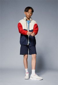 Fargeblokkerte vindjakker i kremfarge, rød og marineblå; shorts med elastisk midje; hvite sneakers med rosa detaljer; glatte teksturer.