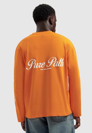 Mann mit dem Rücken zur Kamera, trägt ein leuchtend oranges Langarmshirt mit weißer Aufschrift „Pure Path Own Your Journey“ und blaue Jeans vor einfachem Hintergrund.