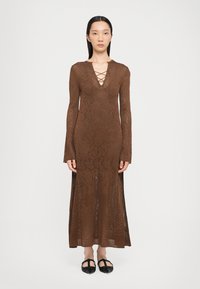 Abito maxi in pizzo marrone con maniche lunghe, scollatura a V profonda e dettagli incrociati. Le fessure laterali mostrano le gambe. Il modello indossa scarpe nere basse.