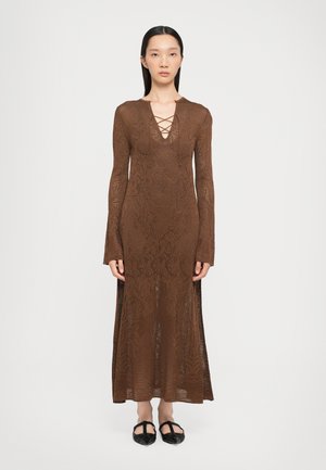 Bruine kanten maxi-jurk met lange mouwen, diepe V-hals en kruiselings detail. Openingen aan de zijkanten tonen de benen. Model draagt zwarte platte schoenen.