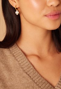 Boucles d'oreilles en or avec un design floral blanc et une texture gaufrée. Portées avec un pull en tricot beige, affichant un décolleté en V.