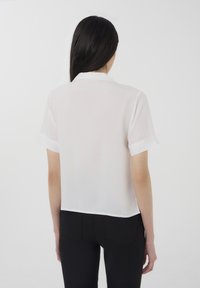 Blusa bianca a maniche corte con design a colletto, texture trasparente e taglio leggermente accorciato. La vista posteriore mostra linee pulite e dettagli minimi.