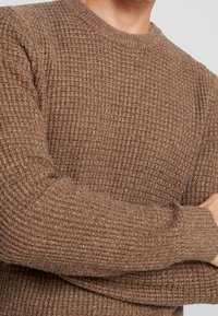 En man i en brun texturerad stickad tröja med långa ärmar står med armarna korsade över bröstet mot en neutral bakgrund.