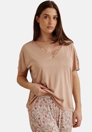Femme portant un haut beige à manches courtes avec des détails en dentelle et un pantalon à motif floral clair, debout devant un fond uni.