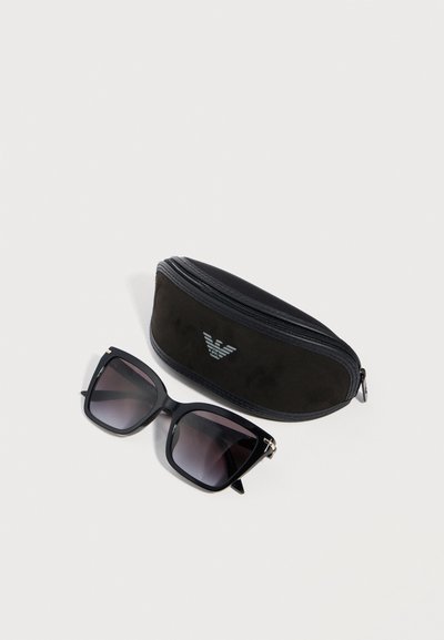Μαύρα γυαλιά ηλίου με τετράγωνο πλαίσιο και σκούρους φακούς με βαθμιαία αλλαγή χρώματος, δίπλα σε μαύρη μαλακή θήκη με φερμουάρ που φέρει ασημί λογότυπο αετού.