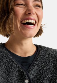 Donkergrijze fleece jas met een gestructureerd oppervlak, met een ronde halslijn en een knoopsluiting, gelaagd over een zwart shirt.