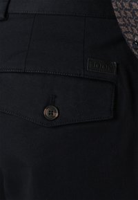 Black textured fabric pant with a back pocket, bronze button, and a label embossed with "JOOP." Stitching is visible along the seams.