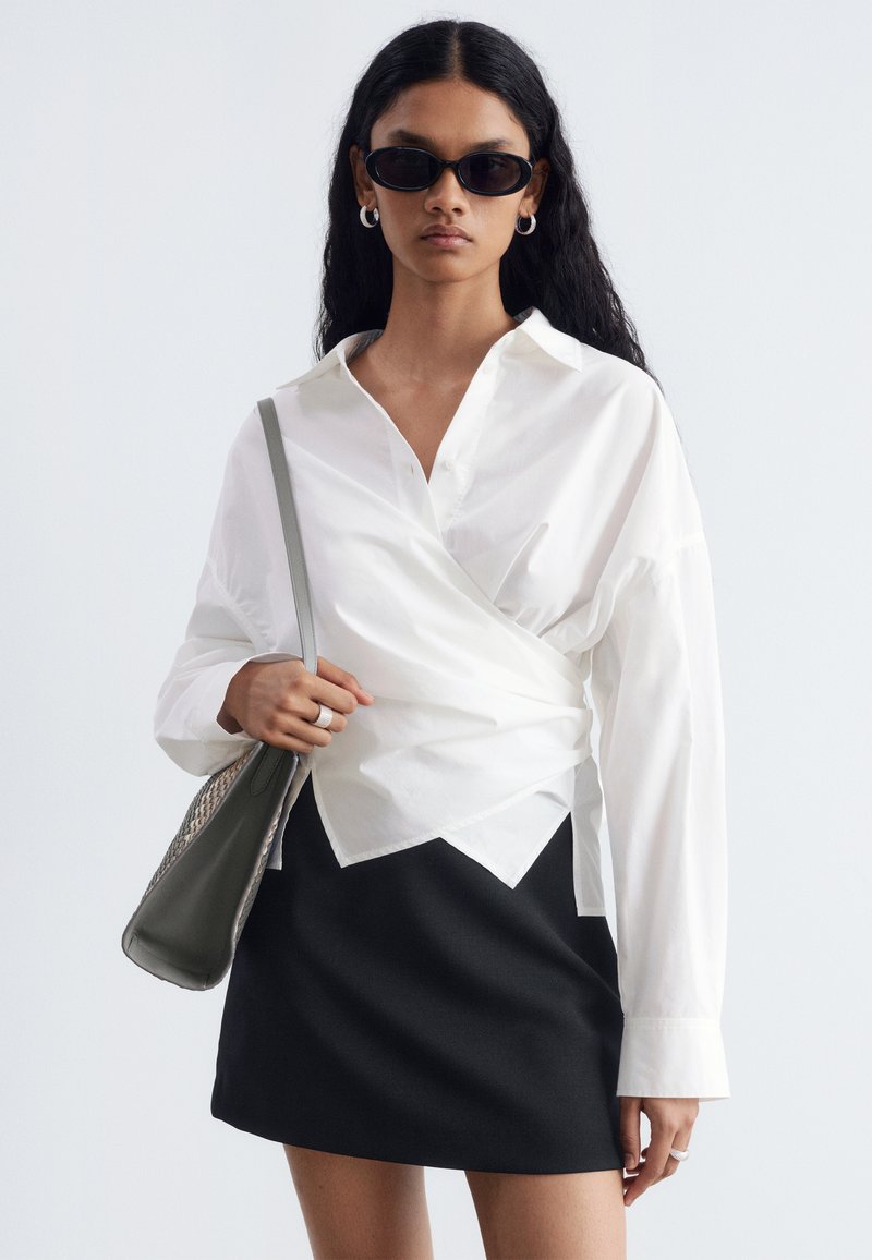 Femme portant des lunettes de soleil noires, un chemisier cache-cœur blanc, une jupe noire, des boucles d'oreilles anneaux argentées et un sac à épaule gris.