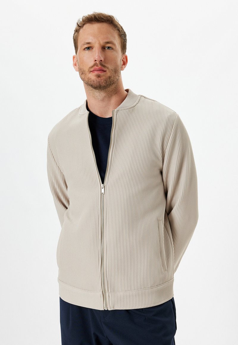 Beige ribbet jakke med lynlås foran, rund halsudskæring og sidelommer. Tekstureret materiale med elastiske manchetter og kant.