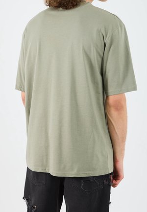 Personne portant un t-shirt à manches courtes vert olive et un jean noir déchiré, vue de dos sur un fond blanc uni.