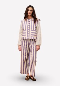 Frau in beige- und lavendelfarbener vertikal gestreifter Weste und Hose, mit einem beigen, strukturierten Oberteil mit Ballonärmeln und dunkelschwarzen flachen Schuhen, steht.