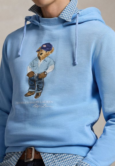 Svijetloplava dukserica s medvjedom koji nosi kapu, traper košulju, traperice i čizme, slojevito odjevena preko plave košulje na kockice.
