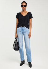 Sieviete melnā V veida kakla T-kreklā, gaiši zilās džinsās, melnās zābakos, saulesbrillēs, turot melnu somu, stāvot pret vienmērīgu gaišu fonu.