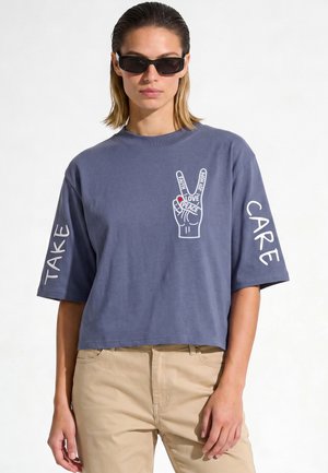 Femme portant des lunettes de soleil foncées et une chemise bleue à manches courtes avec un symbole de paix et les mots "Take Care" imprimés sur les manches.