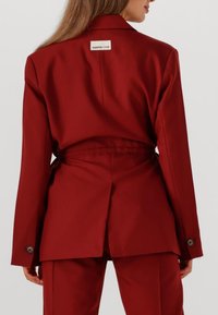 Bordeaux gekleed blazer met een getailleerde taille en dubbele split, voorzien van een label aan de achterkant en knopaccenten op de mouwen.