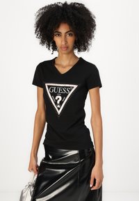 T-shirt nera a V con logo triangolare con accenti argentati e strass, abbinata a una gonna nera lucida.