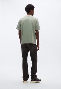 Svetlo zelené tričko s krátkym rukávom, voľný strih, skombinované s tmavohnedými džínsami. Model má na sebe čierno-bežové tenisky s logom.