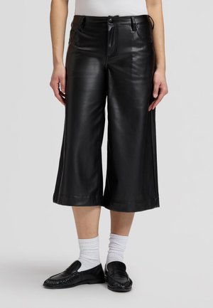 Personne portant des culottes en cuir larges noires, des chaussettes blanches, des mocassins noirs et un haut blanc, debout devant un fond gris clair uni.