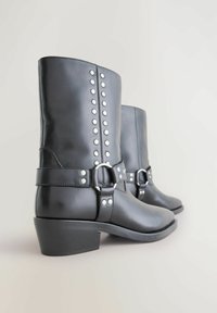 Botas tobilleras de cuero negro con tachuelas plateadas, una correa ancha con hebilla y un tacón bajo y grueso. Textura suave con un acabado pulido.