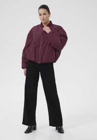 Doudoune bordeaux avec un col haut, une fermeture éclair sur le devant et des poches latérales, associée à un pantalon noir à jambes larges et des bottes à talons noires.