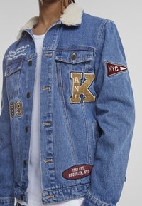 Denim jacket with a shearling collar, blue color, featuring embroidered patches, including letters and a borough emblem. Snap front closure.