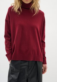 Pull col roulé bordeaux à manches longues en tissu doux avec détails côtelés au niveau du col et de l'ourlet. Associé à un pantalon en cuir noir.