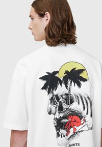 Hvit bomulls-T-skjorte med et bakre motiv av en hodeskalle med palmer, en gul sol og en rød katt. Løs passform og korte ermer.