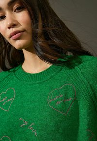 Maglione verde a maglia con scollatura a coste, caratterizzato da cuori ricamati rosa e motivi testuali come "cherie amour" e "my love".
