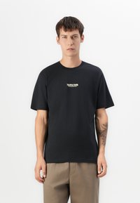 Young man with short dark hair wearing a black printed T-shirt and beige pants, standing against a plain white background.