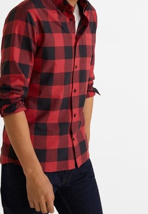 Uomo che indossa una camicia a scacchi rossa e nera con le maniche arrotolate e jeans blu scuro, su uno sfondo bianco semplice.