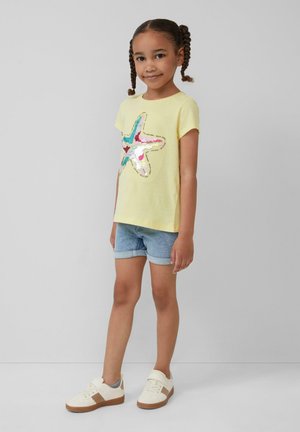 Geel T-shirt met korte mouwen met een kleurrijk sterrensequinsmotief, gecombineerd met blauwe denimshorts en witte sneakers met een bruine zool.