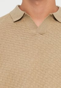 Beżowy dzianinowy polo z fakturowanym wzorem w diamenty, z kołnierzem w kształcie wcięcia i miękkim, lekkim materiałem.