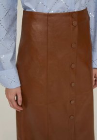 Gonna in finta pelle marrone con forma aderente, caratterizzata da una chiusura a bottoni frontale e una texture liscia. Abbinata a una camicia a righe blu chiaro.