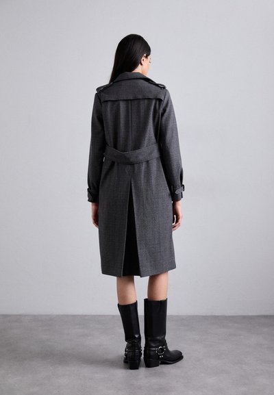 Grau trenchcoat mit Schulterklappen, tailliertem Bund und Rückenschlitz. Getragen mit schwarzen, knielangen Stiefeln mit dekorativen Beschlägen.