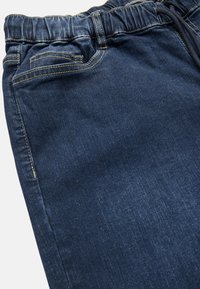 Denimjoggers i mörkblått med elastisk midja, snörning och framficka. Har kontrasterande gul sömnad och en slät yta.