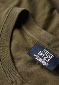T-shirt in cotone verde oliva con scollo rotondo e bordi a costine; presenta un'etichetta blu con testo bianco, "SUPER DRY 23 Athletique."