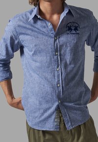 Ljusblå långärmad skjorta med knappar i en texturerad tyg, med en logotyp med två polo-spelare på vänster bröst. Skräddarsydd passform.