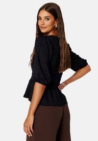 Zwarte blouse met textuur, pofmouwen, peplum taille en een ronde hals, gecombineerd met een paarse broek. Opvallend door de gelaagde stof en het ontwerp.