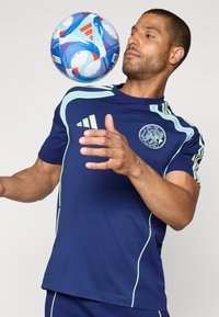 Marine idretts skjorte med lyseblå detaljer og jersey-logo. Holder en blå, rød og hvit mønstrete fotball, med armene strukket ut.