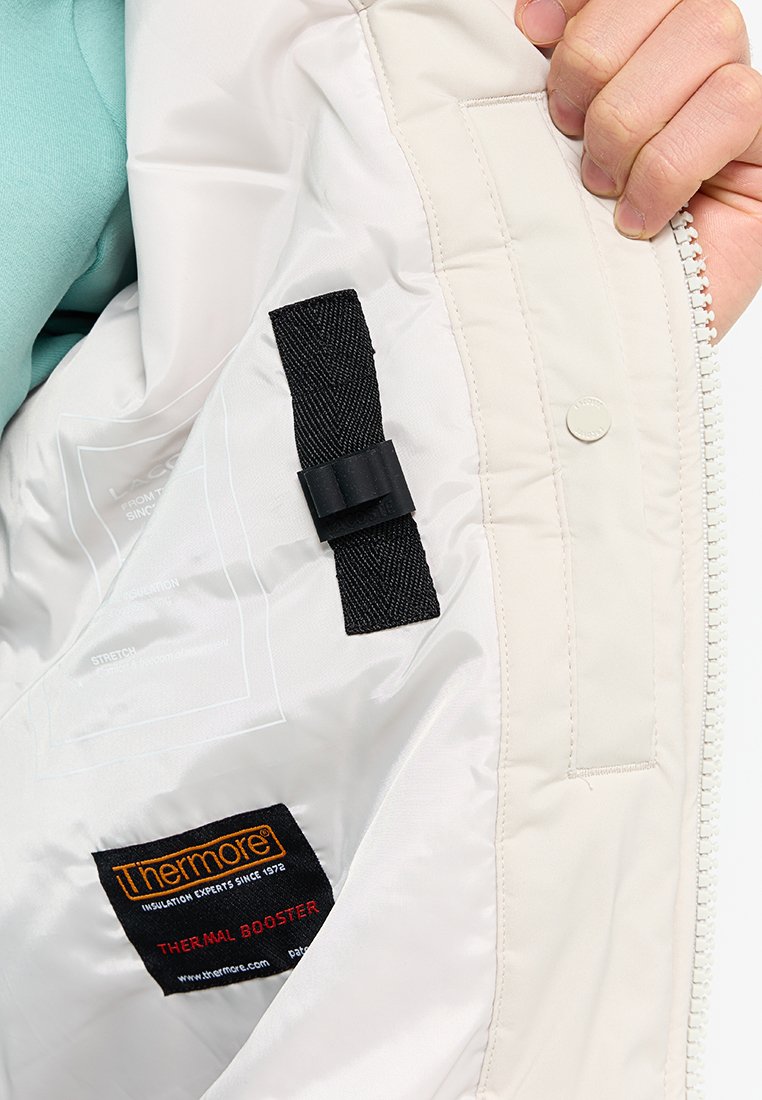 Interno di una giacca isolata bianca che mostra una cinghia nera, etichette con "Thermore" e "Thermal Booster", e una texture trapuntata.