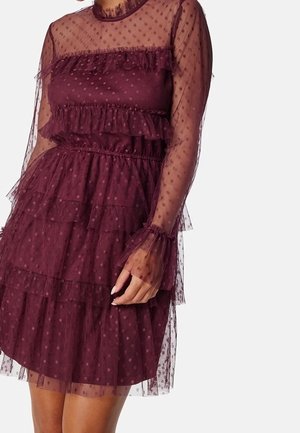 Robe bordeaux transparente à pois avec volants superposés, manches longues et taille cintrée, portée par une personne avec la main posée sur la hanche.