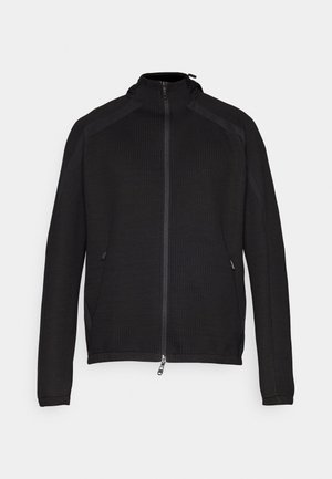 Black zip-up jacket with textured fabric, high collar, and contrasting side panels. Features zipper pockets and a logo on the chest.