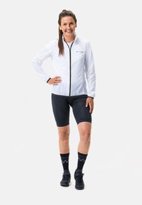 Weiße leichte Jacke mit Reißverschluss, schwarze Radsportshorts und schwarze Socken mit blauen Mustern, kombiniert mit schwarzen Mountainbike-Schuhen.