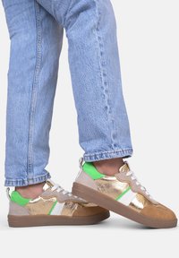 Sneaker con tomaia metallica dorata, dettagli bianchi e verdi, camoscio beige e suola in gomma marrone, abbinate a jeans in denim chiaro.