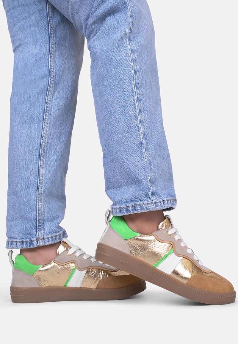 Sneaker con tomaia metallica dorata, dettagli bianchi e verdi, camoscio beige e suola in gomma marrone, abbinate a jeans in denim chiaro.