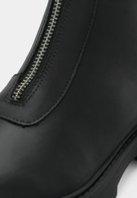Μαύρο δερμάτινο μποτάκι μέχρι τον αστράγαλο με μεταλλική λεπτομέρεια φερμουάρ, λεία υφή, ραφές και στρογγυλή μύτη.