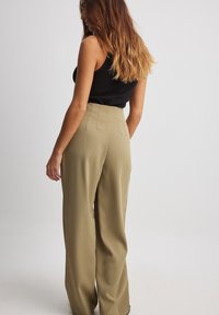 Kalhoty s vysokým pasem a širokými nohavicemi v olivově zelené látce s hladkou texturou a předními záhyby. Nošeny s černým topem.