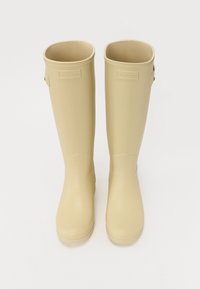 Botas de lluvia de goma beige con acabado mate, diseño alto y puntas redondeadas. Presentan una correa lateral y un detalle de logotipo cerca del borde superior.