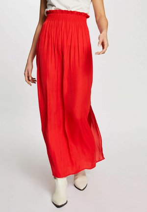Gonna maxi rossa realizzata in un tessuto leggero e fluido con una vita elastica arricciata. Texture liscia, spacco laterale fino al ginocchio, abbinata a stivaletti bianchi.